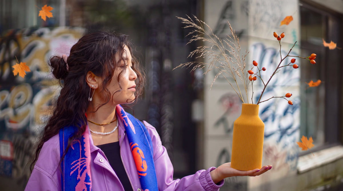 Frau in lila Jacke hält die gerillte Vase Flasche von dennismaass. in Ocker mit Trockengras und Hagebutten vor einer herbstlichen Graffitiwand