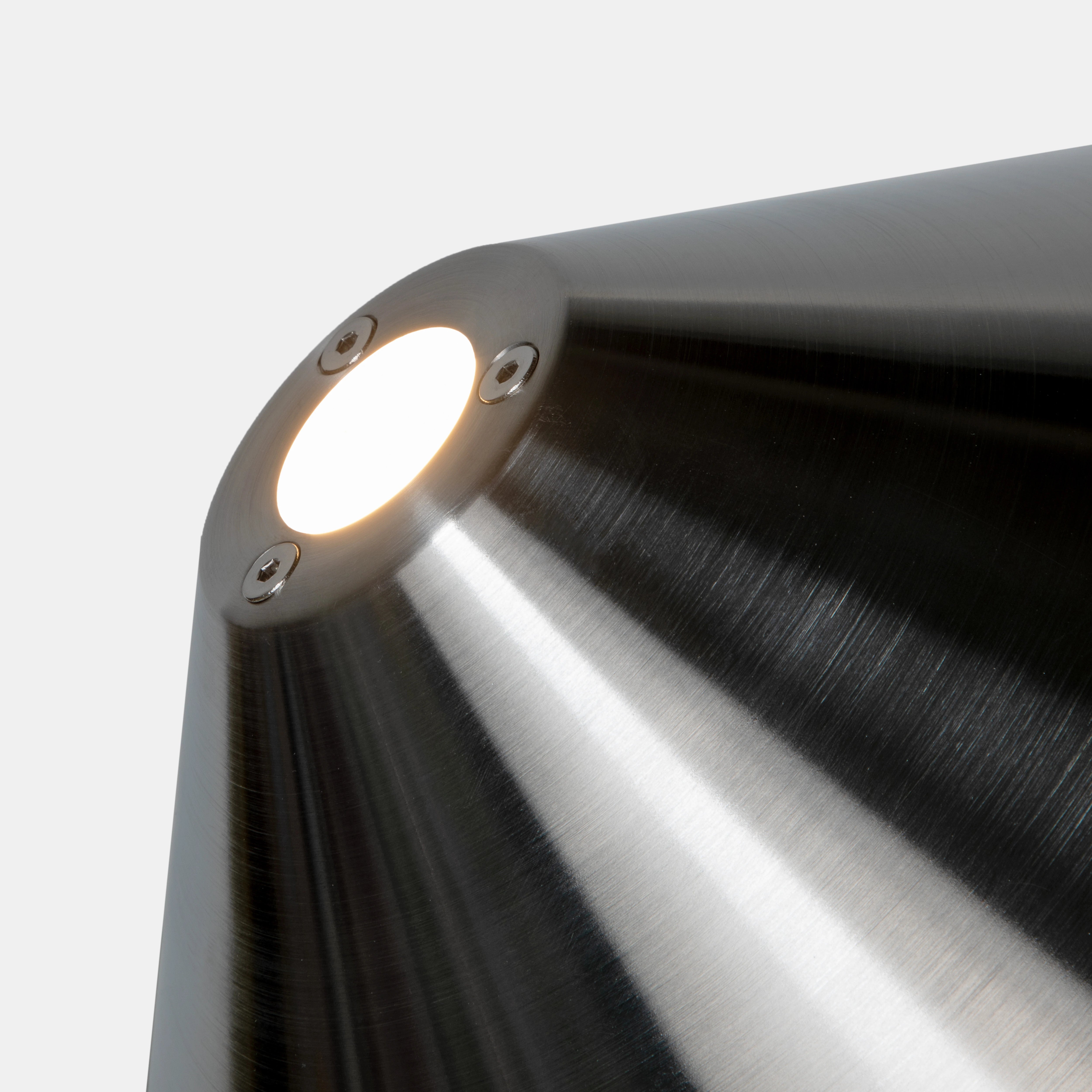 Detail der oberen Aussparung im Schirm: Schraubentrio und warmweißes Licht des E14-LED-Leuchtmittels vor gebürstetem Edelstahl.